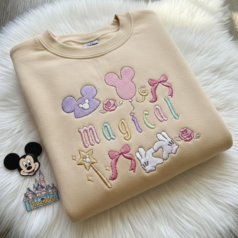 May include: Beige sweatshirt with embroidered "Magical" design. Features Mickey Mouse head shapes, a pink balloon, bows, a wand, and Mickey Mouse gloves. Includes a Mickey Mouse pin and Disneyland pin.