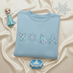 May include: A light blue sweatshirt with embroidered designs of gloves, a snowflake, a dress, and a castle. A small doll and a decorative box are also in the image, along with a large snowflake.