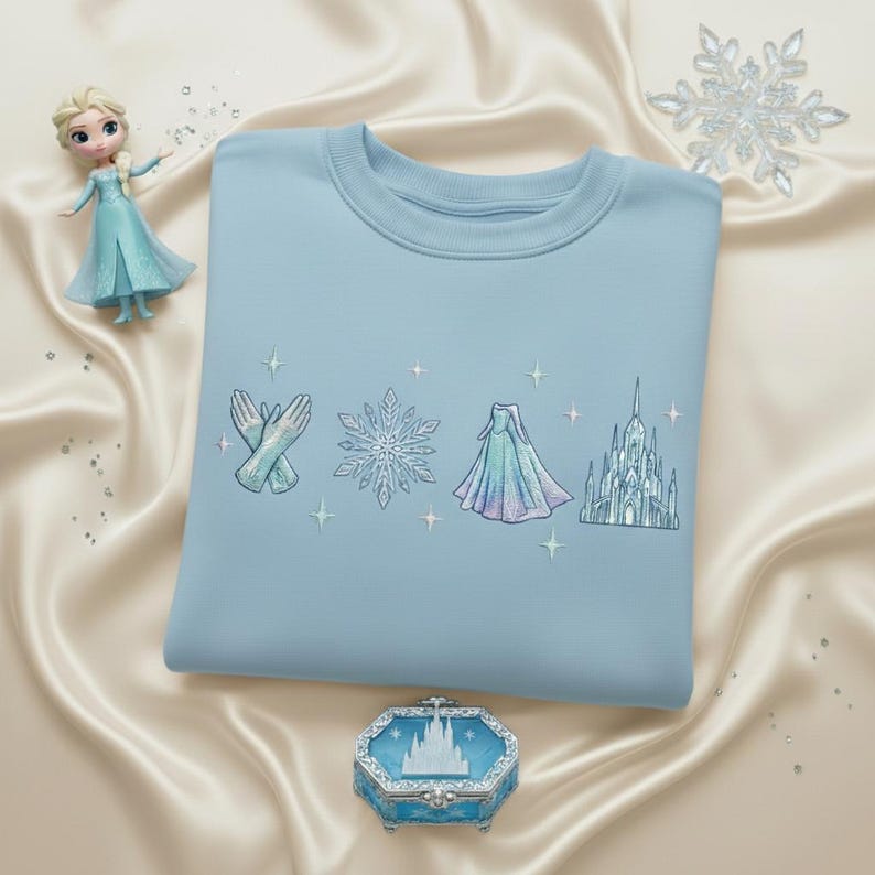May include: A light blue sweatshirt with embroidered designs of gloves, a snowflake, a dress, and a castle. A small doll and a decorative box are also in the image, along with a large snowflake.