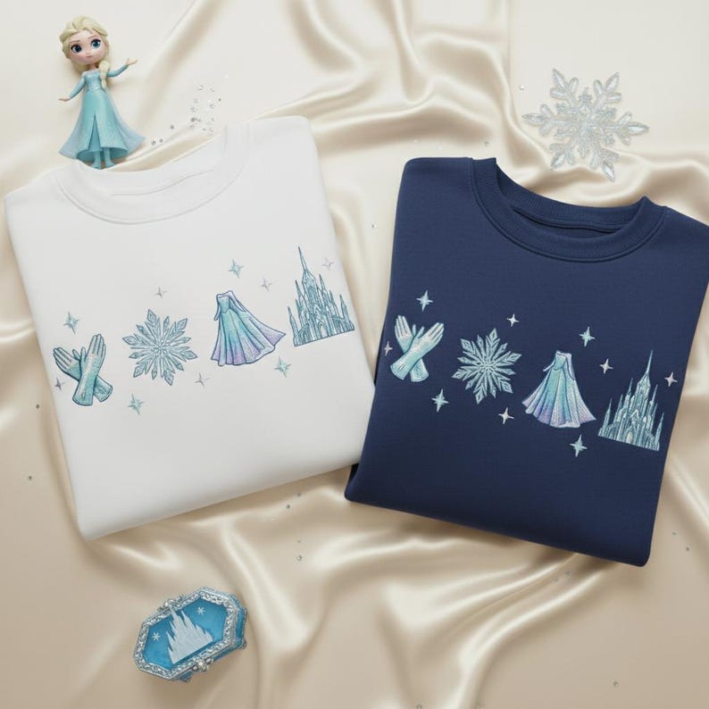 May include: Two sweatshirts, one white and one navy blue, with embroidered designs of a snowflake, gloves, a cape, and a castle. A small doll and a decorative box are also in the image. The sweatshirts are on a satin surface.