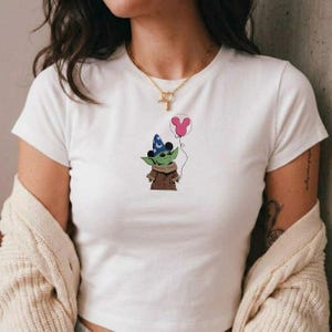 Puede incluir: Camiseta blanca con un personaje de dibujos animados de piel verde, que lleva un sombrero de mago azul y una capa marr&oacute;n, sosteniendo un globo rosa de Mickey Mouse. El dise&ntilde;o est&aacute; centrado en la camiseta.