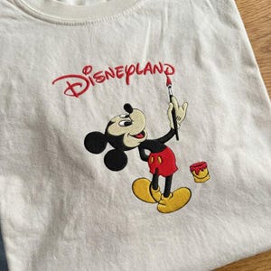 Pu&ograve; includere: T-shirt color crema con ricamo di Mickey Mouse che tiene un pennello, con la scritta "Disneyland" in rosso sopra. Mickey indossa pantaloncini rossi e scarpe gialle. Un piccolo barattolo di vernice &egrave; accanto a lui.