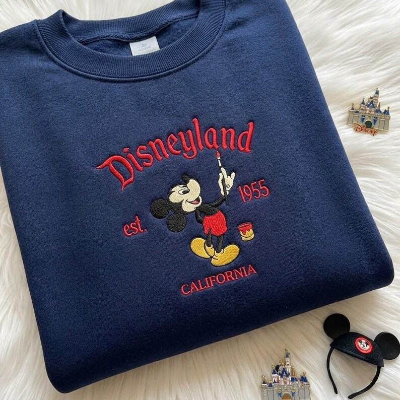 May include: A navy blue crewneck sweatshirt featuring red and white embroidery of Mickey Mouse holding a paintbrush. The text reads Disneyland, est. 1955, and CALIFORNIA. The design includes a small paint bucket and a castle pin nearby.