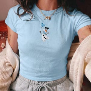May include: Light blue short-sleeved t-shirt featuring an embroidered Olaf design holding a Mickey Mouse balloon. The shirt is a solid light blue color and has a crew neck. The design is in white, black, and orange.
