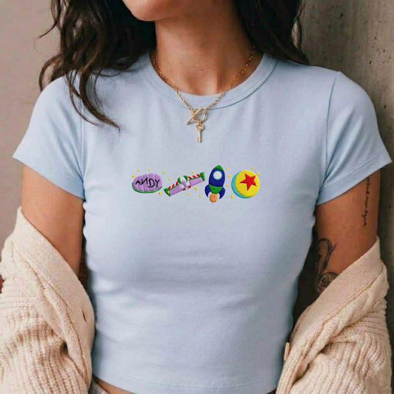 May include: Light blue crop top with embroidered graphics. The design includes the word "ANDY", a purple and green candy, a blue and green rocket, and a yellow ball with a red star. The shirt has short sleeves and a crew neck.