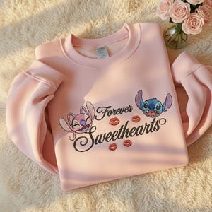 May include: A light pink sweatshirt featuring the characters Angel and Stitch from Lilo & Stitch. The sweatshirt has the words "Forever Sweethearts" with kiss graphics. The sleeves are long and the material appears soft.