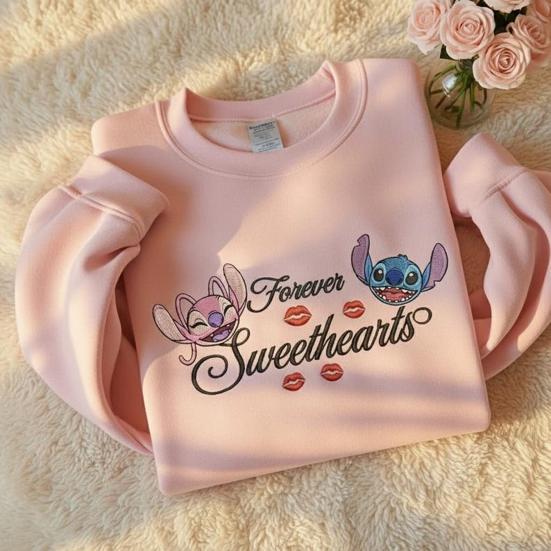 May include: A light pink sweatshirt featuring the characters Angel and Stitch from Lilo & Stitch. The sweatshirt has the words "Forever Sweethearts" with kiss graphics. The sleeves are long and the material appears soft.
