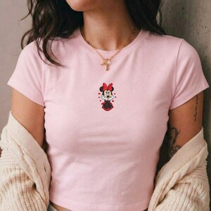 May include: A light pink t-shirt featuring a Minnie Mouse graphic. The graphic shows Minnie Mouse in a red dress and bow, surrounded by hearts. The shirt has short sleeves and a crew neck.