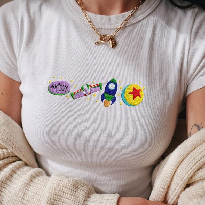 May include: White t-shirt with embroidered Toy Story-themed designs. The design includes the word "ANDY" in a purple oval, a green and purple spaceship, a blue and yellow rocket, and a yellow ball with a red star.