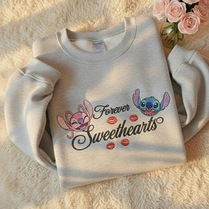 May include: A light gray sweatshirt featuring the characters Angel and Stitch with the words "Forever Sweethearts" and red kiss graphics. The sweatshirt has a crew neck and long sleeves.