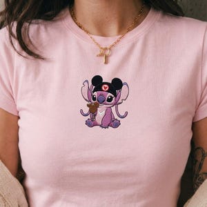 May include: A light pink t-shirt featuring an embroidered cartoon character in a nurse's cap holding a small brown object. The character is purple with black ears and a red heart on the cap. A gold necklace is visible.