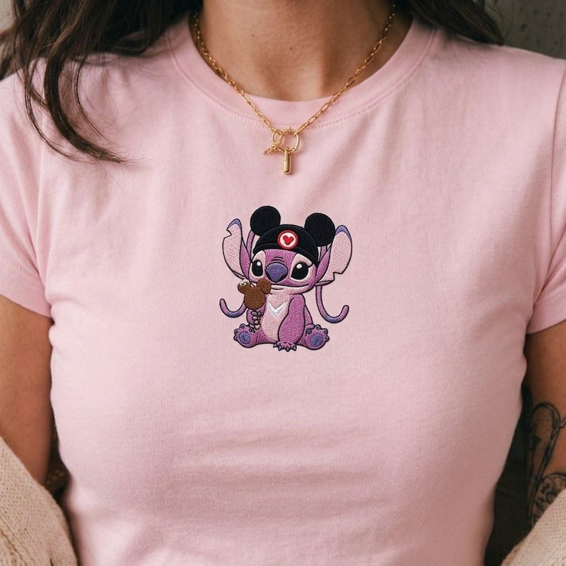 May include: A light pink t-shirt featuring an embroidered cartoon character in a nurse's cap holding a small brown object. The character is purple with black ears and a red heart on the cap. A gold necklace is visible.