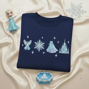 May include: Navy blue sweatshirt featuring embroidered elements from a popular animated film. The design includes gloves, a snowflake, a cape, and a castle, with star accents. A figurine and a decorative box with a castle design are also present.