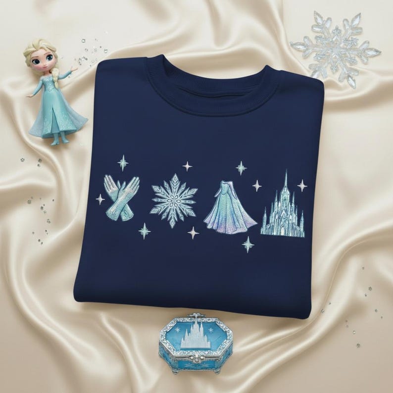 May include: Navy blue sweatshirt featuring embroidered elements from a popular animated film. The design includes gloves, a snowflake, a cape, and a castle, with star accents. A figurine and a decorative box with a castle design are also present.