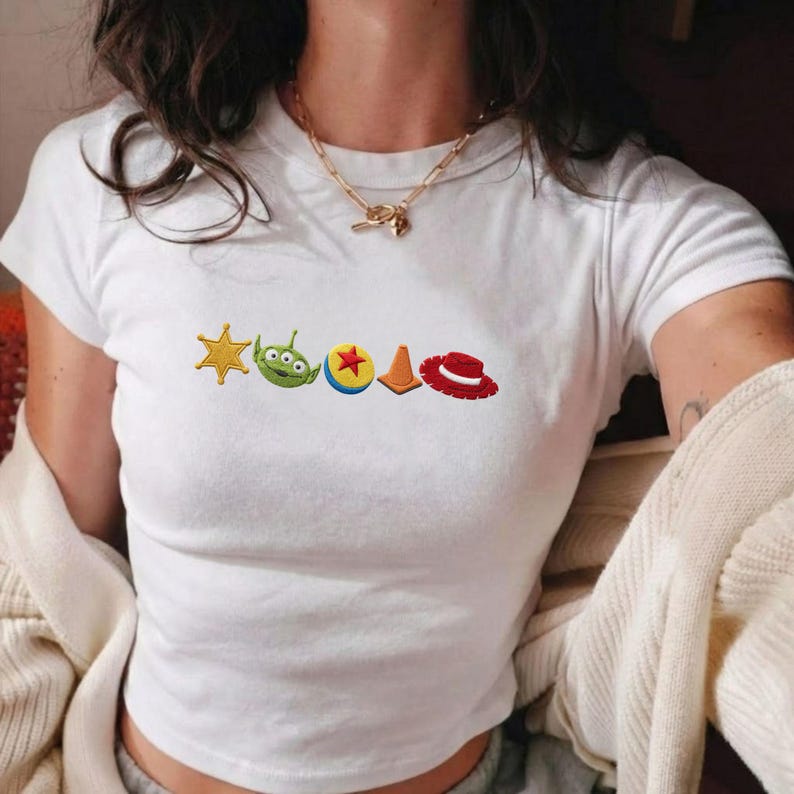 K&ouml;nnte beinhalten: Ein wei&szlig;es T-Shirt mit kurzen &Auml;rmeln und einer horizontalen Reihe aus f&uuml;nf gestickten Symbolen: ein gelber Sheriffstern, ein gr&uuml;ner dreiaugiger Au&szlig;erirdischer, ein gelb-roter Ball, ein orangefarbener Verkehrskegel und ein roter Cowboyhut.