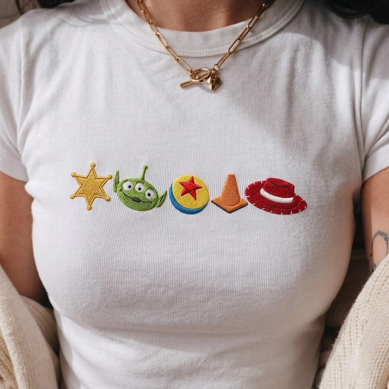 K&ouml;nnte beinhalten: Ein wei&szlig;es kurz&auml;rmeliges T-Shirt mit einer horizontalen Reihe aus f&uuml;nf gestickten Symbolen: einem gelben Sheriffstern, einem gr&uuml;nen dreiaugigen Alienkopf, einem gelben Ball mit rotem Stern, einem orangefarbenen Verkehrskegel und einem roten Cowboyhut mit wei&szlig;em Band.