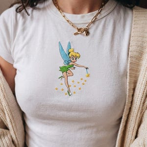 Puede incluir: Una camiseta blanca de manga corta con un bordado central de una hada rubia con alas azules, vestida con un vestido verde y sosteniendo una varita que emite pequeñas estrellas amarillas. Un collar de cadena dorada descansa en el cuello.