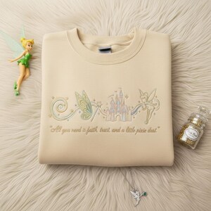 May include: A cream-coloured sweatshirt with an embroidered design of a fairy, castle, and the text "All you need is faith, trust, and a little pixie dust." A fairy figurine, glitter jar, and necklace are also in the image.