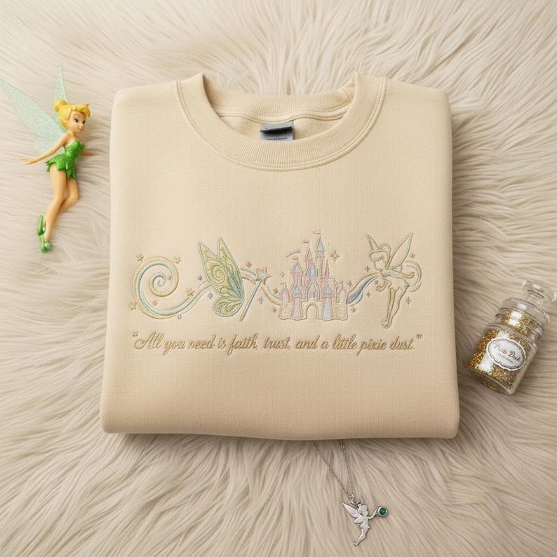 May include: A cream-coloured sweatshirt with an embroidered design of a fairy, castle, and the text "All you need is faith, trust, and a little pixie dust." A fairy figurine, glitter jar, and necklace are also in the image.