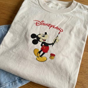 Pu&ograve; includere: T-shirt color crema con un ricamo di Topolino. Topolino &egrave; raffigurato con un pennello, con la scritta "Disneyland" ricamata in rosso sopra. La maglietta ha un girocollo classico ed &egrave; abbinata a jeans blu.