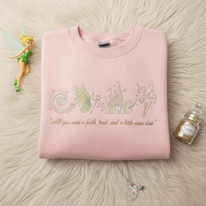 May include: A pale pink sweatshirt with an embroidered design of a castle, fairy wings, and a fairy. The text "All you need is faith, trust, and a little pixie dust" is below the design. A fairy figurine and glitter jar are also in the image.