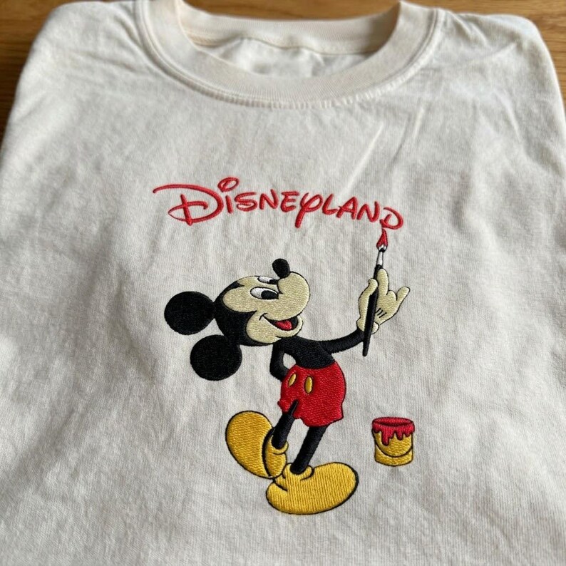 Pu&ograve; includere: T-shirt color crema con un ricamo di Topolino. Mickey &egrave; raffigurato con un pennello, con la scritta "Disneyland" in rosso sopra. Un piccolo barattolo di vernice &egrave; incluso nel design.