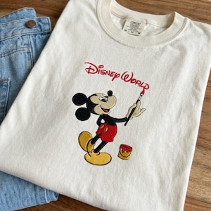 K&ouml;nnte beinhalten: Cremefarbenes T-Shirt mit gesticktem Mickey Mouse, der "Disney World" in Rot malt. Das Shirt hat einen Rundhalsausschnitt und kurze &Auml;rmel. Eine blaue Jeans ist im Hintergrund zu sehen.