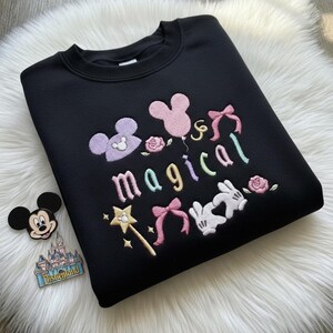 May include: Black sweatshirt with colorful embroidered design. Features the word "magical" with Mickey Mouse and Minnie Mouse imagery, bows, a wand, and roses. A Mickey Mouse pin and a Disneyland pin are also visible.