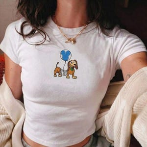 Op de afbeelding: Wit T-shirt met een cartoon teckel die een blauwe ballon vasthoudt. De hond heeft een veerkrachtig lichaam en is bruin en beige. Het shirt heeft een ronde hals en korte mouwen.