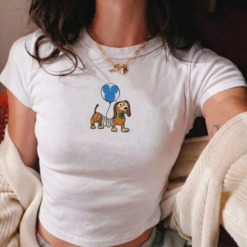 Op de afbeelding: Wit T-shirt met een cartoon teckel die een blauwe ballon vasthoudt. De hond heeft een veerkrachtig lichaam en is bruin en beige. Het shirt heeft een ronde hals en korte mouwen.