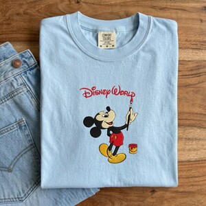 K&ouml;nnte beinhalten: Hellblaues T-Shirt mit gesticktem Mickey Mouse-Design, der einen Pinsel h&auml;lt, und dem Schriftzug "Disney World" in roter Schrift. Das T-Shirt ist gefaltet, mit einer Jeans auf der linken Seite. Das T-Shirt hat einen Rundhalsausschnitt und ein Etikett "Comfort Colors".
