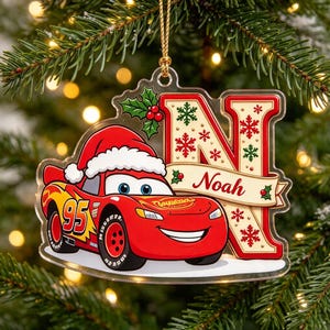 Può includere: Un addobbo natalizio in acrilico trasparente con un'auto da corsa rossa che indossa un cappello da Babbo Natale accanto a una grande lettera N decorata con fiocchi di neve rossi e verdi. Un nastro riporta il nome Noah in rosso.