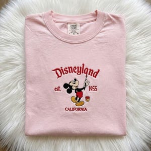 May include: A light pink short-sleeved t-shirt featuring an embroidered graphic of Mickey Mouse holding a paintbrush. The text reads Disneyland, est. 1955, and CALIFORNIA in red thread. The shirt has a Comfort Colors brand label at the neck.