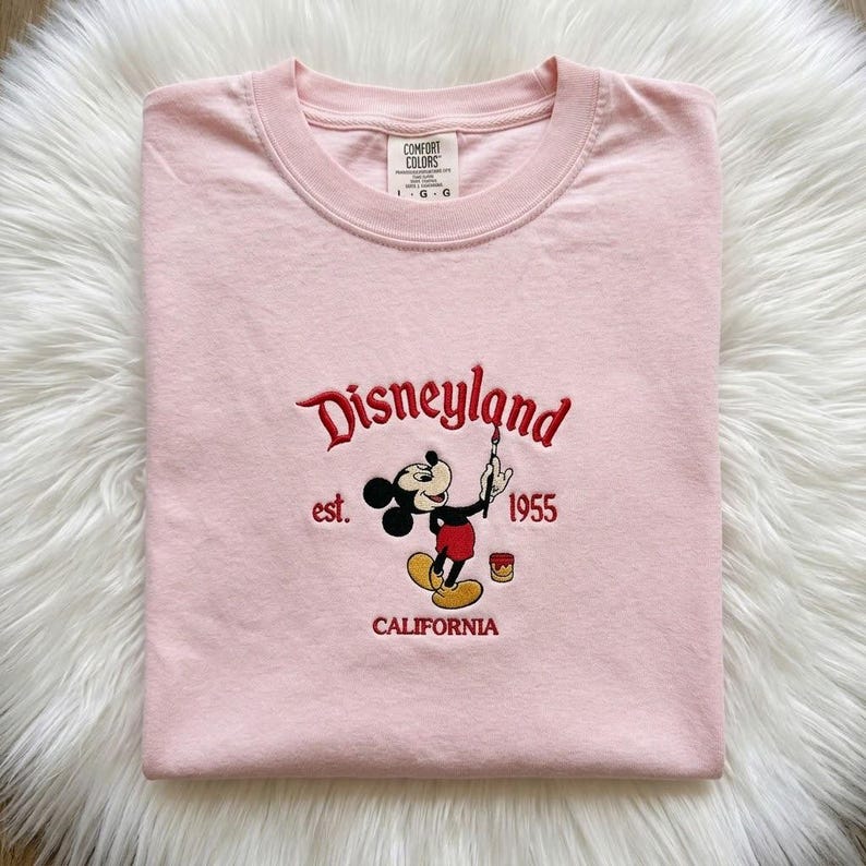May include: A light pink short-sleeved t-shirt featuring an embroidered graphic of Mickey Mouse holding a paintbrush. The text reads Disneyland, est. 1955, and CALIFORNIA in red thread. The shirt has a Comfort Colors brand label at the neck.