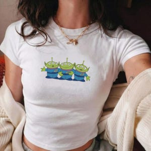 May include: A white short-sleeved t-shirt featuring a graphic print of three green three-eyed aliens wearing blue uniforms. The aliens are positioned side-by-side in the center of the chest. The shirt has a crew neckline and a fitted silhouette.