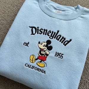 May include: A light blue crewneck sweatshirt featuring black embroidered text that reads Disneyland est. 1955 CALIFORNIA. Centered is an embroidered graphic of Mickey Mouse wearing red shorts and yellow shoes, positioned between the text elements.