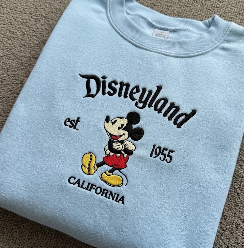 May include: A light blue crewneck sweatshirt featuring black embroidered text that reads Disneyland est. 1955 CALIFORNIA. Centered is an embroidered graphic of Mickey Mouse wearing red shorts and yellow shoes, positioned between the text elements.