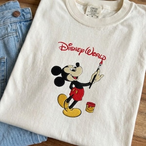 K&ouml;nnte beinhalten: Cremefarbenes T-Shirt mit einem gestickten Mickey Mouse-Design. Mickey wird mit einem Pinsel dargestellt, dar&uuml;ber die Worte "Disney World" gestickt. Das Shirt wird mit blauen Jeans kombiniert.