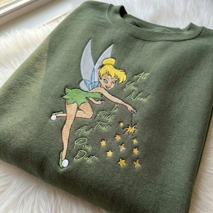 Peut inclure: Sweat-shirt vert olive avec une broderie de la fée Clochette. Le design comprend le texte « All You Need Is Faith, Trust, And Pixie Dust » et des étoiles jaunes. La fée est représentée avec des ailes détaillées et une tenue verte.
