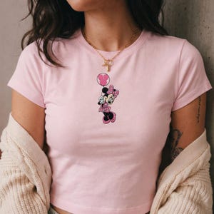 May include: A light pink t-shirt featuring a Minnie Mouse graphic. The graphic shows Minnie Mouse in a pink polka-dot dress and bow, holding a pink balloon. The shirt has short sleeves and a crew neck.