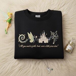 May include: Black sweatshirt with an embroidered design of a castle, fairy wings, and a fairy. The text "All you need is faith, trust, and a little pixie dust" is below the design. A fairy figurine and a glitter jar are also in the image.