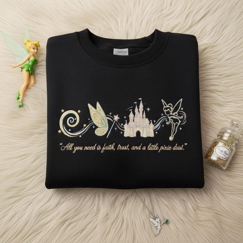 May include: Black sweatshirt with an embroidered design of a castle, fairy wings, and a fairy. The text "All you need is faith, trust, and a little pixie dust" is below the design. A fairy figurine and a glitter jar are also in the image.