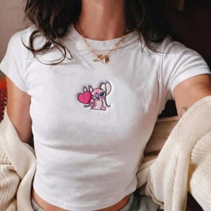 May include: White crop top with a small embroidered design of a pink cartoon character holding a pink heart. The character has one eye and large ears. The shirt has short sleeves.