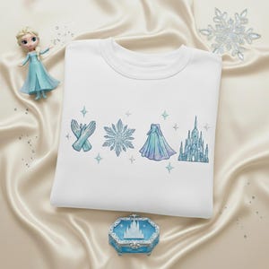 May include: White crewneck sweatshirt with embroidered light blue gloves, snowflake, dress, and castle designs. A small doll and decorative box with a castle design are also present. The sweatshirt is on a satin surface.