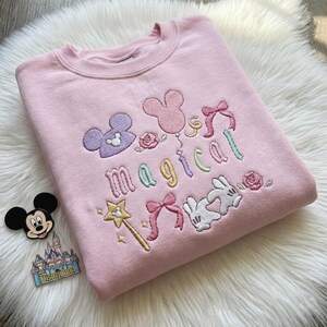 May include: A light pink sweatshirt with embroidered Disney-themed graphics. The design includes a Mickey Mouse head, a wand, bows, and the word "magical" in pastel colors. A Mickey Mouse pin and Disneyland pin are visible.