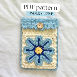 May include: A crochet Kindle sleeve featuring a cream-colored base with a large blue flower motif. The sleeve has a light blue scalloped flap secured by a wooden button. Text above reads PDF pattern KINDLE SLEEVE. The design is rectangular and textured.
