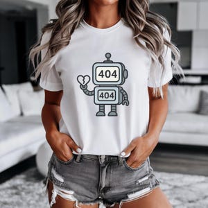May include: White t-shirt featuring a cartoon robot design with the text "404" on its head and body. The robot is holding a broken heart. The design is in shades of gray, black, and red.