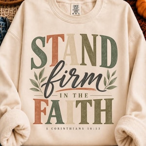 Puede incluir: Una sudadera de cuello redondo color crema con el texto STAND firm IN THE FAITH y 1 CORINTHIANS 16:13. El diseño utiliza tonos apagados de terracota, verde salvia y crema con ilustraciones de hojas a los lados de la palabra firm.