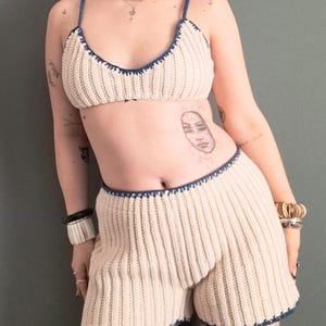 May include: Cream-colored crochet two-piece set with navy blue trim. The set includes a bra top with thin straps and high-waisted shorts. The crochet pattern features vertical ribbing. The edges of the bra and shorts are finished with a scalloped navy blue trim.