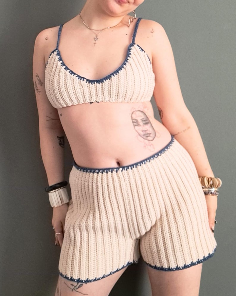 May include: Cream-colored crochet two-piece set with navy blue trim. The set includes a bra top with thin straps and high-waisted shorts. The crochet pattern features vertical ribbing. The edges of the bra and shorts are finished with a scalloped navy blue trim.